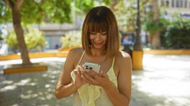 Güneşli bir günde şehir parkında akıllı telefon kullanan kısa esmer saçlı güzel bir genç kadın.