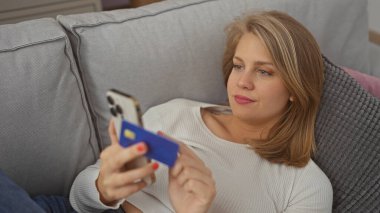 Kapalı bir koltukta oturan sarışın kadın elinde akıllı telefon ve kredi kartıyla internetten alışveriş yapmayı düşünüyor..