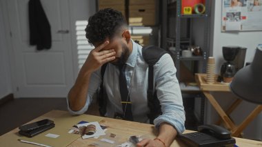 Stressed hispanic man sitting in a detective's office with disarrayed evidence like a passport and smartphone. clipart