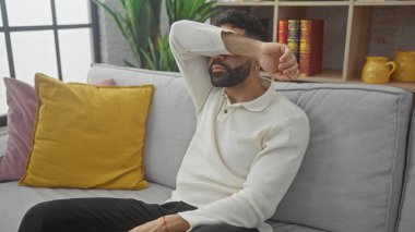 Evdeki gri bir kanepede oturan yorgun bir adam, koluyla yüzünü kapatıyor, yorgunluk ve hayal kırıklığını gösteriyor..