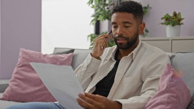 Afrikalı Amerikalı bir adam modern bir oturma odasında telefonla bir belgeyi tartışıyor..