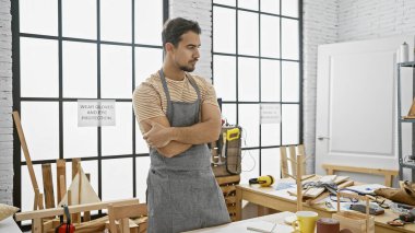 Kollarını kavuşturmuş düşünceli İspanyol bir adam marangozluk atölyesinde duruyor. Etrafı alet ve ahşap işçiliğiyle çevrili..