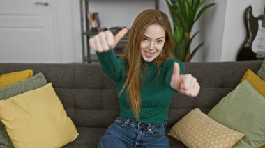 Kafkasyalı genç bir kadın, rahat bir oturma odasındaki kanepede otururken baş parmağını kaldırıyor..