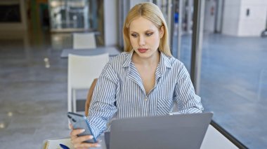 Modern şehir içinde dizüstü bilgisayarda çalışan sarışın kadın elinde akıllı telefon, iş kıyafetleri var..