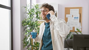 Modern ofiste telefonla konuşan gülümseyen bir adam elinde bir kahve fincanı, arkasında bilgisayar ve kapalı alanda bitkiler olan bir adam..