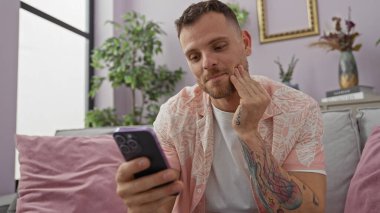 Modern oturma odasındaki koltukta akıllı telefon kullanan sakallı İspanyol bir adam.