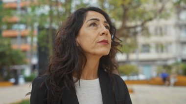 Düşünceli orta yaşlı kadın şehir parkı ortamında yukarı bakıyor, düşüncelere dalmış..