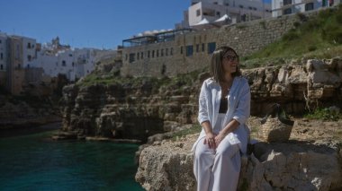 Polignano 'da bir uçurumun kenarında oturan genç bir İspanyol kadın güneşli bir günde kumsalın ve denizin tadını çıkaran bir kısrak, puglia, İtalya..