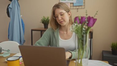 Odaklanmış sarışın bir kadın, yakınlarda bitkileri ve fincanı olan rahat bir ofiste dizüstü bilgisayarla çalışıyor..