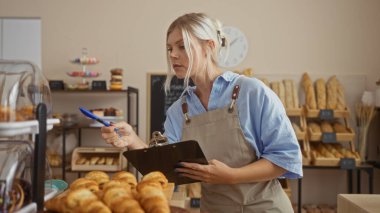 Fırındaki sarışın kadın elinde pano ve kalemle envanterini kontrol ediyor, etrafı ekmek ve pastalarla çevrili, mavi gömlekli ve önlüklü, kapalı bir dükkanda ekmek raflarıyla..