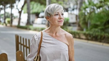 Kısa gri saçlı, olgun bir İspanyol kadın. Düşünceli bir şekilde yeşil bir caddeye bakıyor..