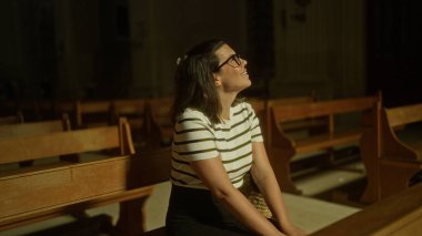 Çizgili tişörtlü genç İspanyol kadın ahşap banklarla İtalyan kilisesinde oturuyor, tefekkür içinde yukarıya bakıyor, dingin bir Hıristiyan atmosferiyle çevrili..