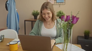 Evde dizüstü bilgisayarda çalışan gülümseyen genç bir kadın. Etrafı çiçeklerle çevrili ve içi rahat..