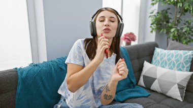 A relaxed young woman enjoys music on headphones while sitting on a blue sofa in a modern living room and applying red lipstick. clipart