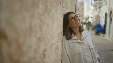 Beautiful young hispanic woman smiling in the charming streets of polignano a mare, puglia, italy, leaning against a stone wall on a sunny day. clipart