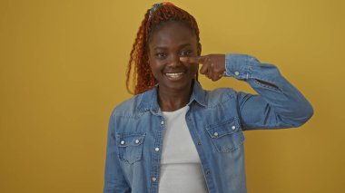 Kot pantolon giymiş, gülümseyen genç bir siyah kadın kendini parlak sarı bir arka plana işaret ediyor.