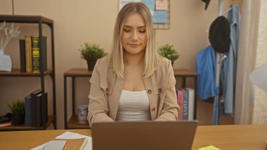 Kitaplar ve bitkilerle çevrili, rahat bir ofis ortamında çalışan çekici sarışın kadın, iyi organize edilmiş bir oturma odasında dizüstü bilgisayarına odaklanıyor..