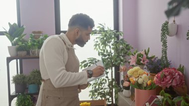 Önlük takan sakallı genç bir adam renkli çiçeklerle dolu bir çiçekçide notlar yazıyor..