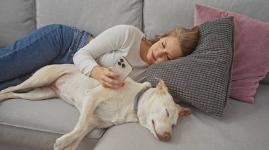 Bir kadın kanepede rahat bir oturma odasında uyuyan bir köpeğe sarılıyor, elinde akıllı bir telefon tutuyor..