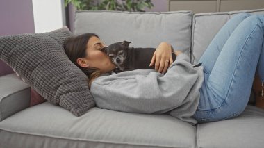 Genç bir İspanyol kadın evcil köpeğini sıcak bir oturma odasında kanepede öpüyor..