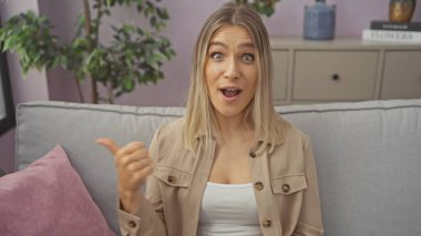 Bej ceketli, çekici, beyaz bir kadın rahat bir oturma odasında oturuyor, ağzı açık ve onu işaret ederek şaşkınlığını anlatıyor..