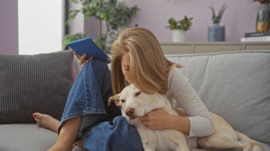 Genç bir kadın sıcak bir oturma odasında tablet tutarken kanepede köpeğine sarılıyor..