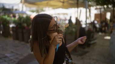 Genç İspanyol kadın, Puglia, İtalya 'daki yaşlı bir kasabanın büyüleyici sokaklarında dondurma yiyor. Etrafı canlı kafeler ve yeşilliklerle çevrili. Güzel bir yaz anını yakalıyor..