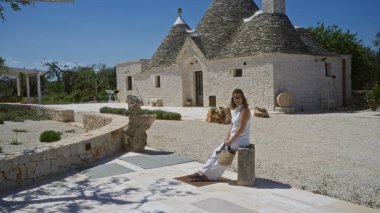 Güzel, genç, İspanyol bir kadın Alberobello 'da geleneksel bir trulli evinin önünde oturuyor. Puglia bölgesinde güneşli bir günün tadını çıkarıyor..