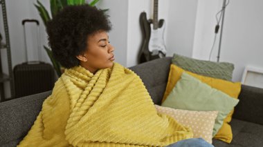 Rahat bir Afrikalı Amerikalı kadın, evinde bir kanepede oturuyor, sarı bir battaniyeye sarılmış, düşünceli bir ifadeyle..