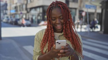 Gülümseyen Afrikalı Amerikalı kadın güneşli bir caddede akıllı telefon kullanıyor.