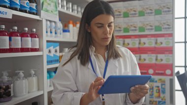 İyi stoklanmış bir eczanede beyaz önlüklü, tablet kullanan odaklanmış bir kadın eczacı..