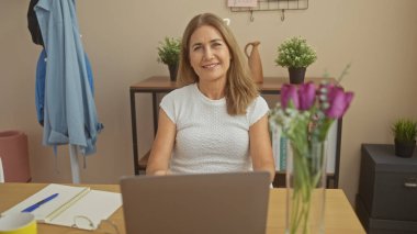 Gülümseyen olgun bir kadın, evinde dizüstü bilgisayarının başında bitki ve dekorla çalışıyor..