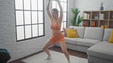 Spor kıyafetli genç bir kadın, geniş bir pencere ve kanepesi olan modern bir oturma odasında yoga yapıyor..