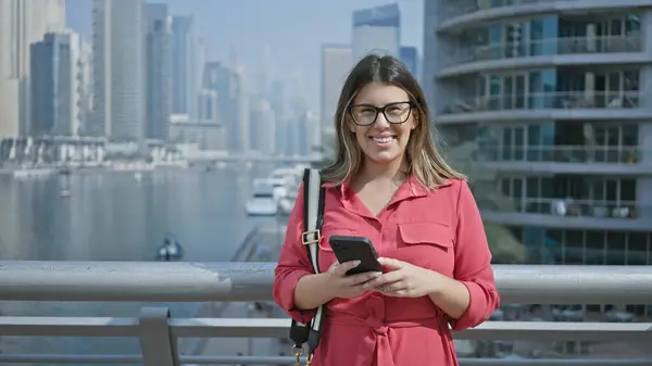 stock image A smiling young adult woman in a red shirt using a smartphone in front of the dubai marina skyline