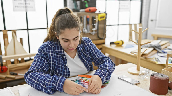 Латиноамериканская молодая женщина внимательно изучает цветные образцы в своей столярной мастерской в помещении.