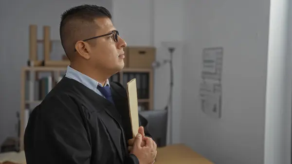 stock image Hispanic man in a formal robe standing in an office, holding a folder with a focused expression, suggesting a legal professional such as a lawyer or judge in a courtroom setting.