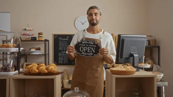 stock image Young man with beard standing in bakery shop holding open sign wearing apron surrounded by pastries and bakery goods