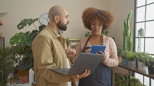 stock image A man and a woman discuss business on a tablet and laptop amid vibrant houseplants inside a sunny flower shop.