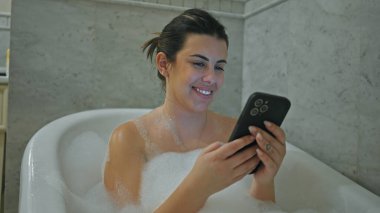 Gülümseyen genç bir kadın modern fayanslı bir banyoda akıllı telefonunu kullanırken köpük banyosundan zevk alır..