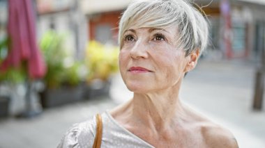 Açık hava ortamında kısa gri saçlı olgun bir İspanyol kadın güzelliği ve düşünceliliği resmediyor.