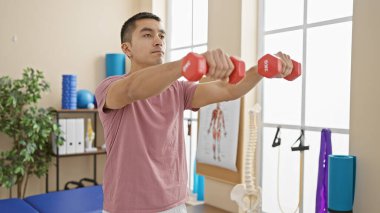 Genç İspanyol adam, parlak bir fizyoterapi kliniğinde dambıllarla egzersiz yapıyor..