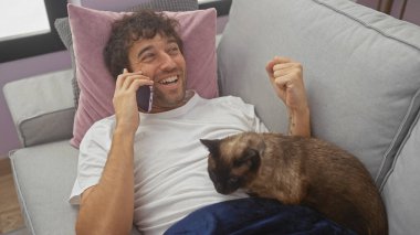 Mutlu bir İspanyol adam, kedisiyle birlikte rahat bir apartman dairesinde kanepede uzanırken telefonda konuşur.