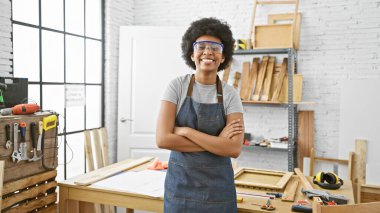 Güvenilir gözlüklü ve kot önlüklü kendinden emin bir Afrikalı kadın iyi aydınlatılmış bir marangozluk atölyesinde kollarını kavuşturuyor..