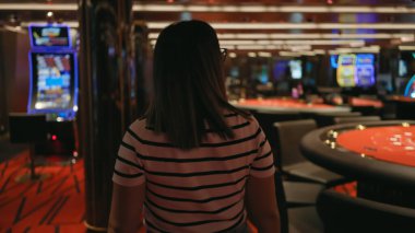 Kumarhanede kumar masaları ve kumar makineleri olan esmer bir kadının arka görüntüsü..