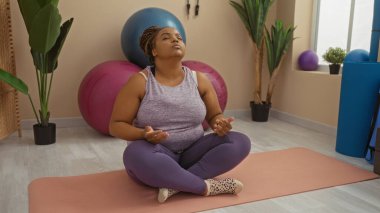Egzersiz topları ve arka planda bitkilerle yoga minderi üzerinde meditasyon yapan bir kadın..