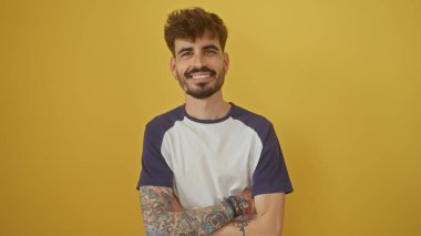 Dövmeli yakışıklı İspanyol adam sarı arka planda kollarını kavuştururken özgüvenle gülümsüyor.
