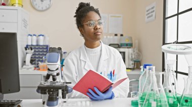 Laboratuvar önlüklü Afrikalı Amerikalı kadın bilim adamı bir laboratuarda mikroskop ve camla bir defter tutuyor..