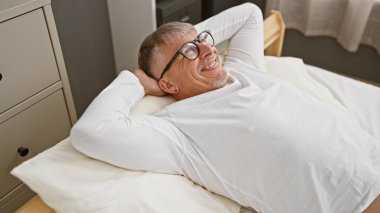 Orta yaşlı bir adam, parlak bir yatak odasında rahatlıyor, rahat bir yatakta mutluluk ve rahatlık yayıyor..