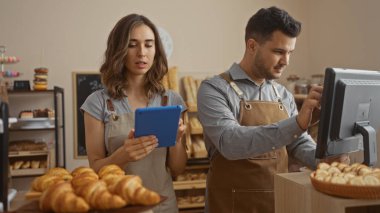 Woman and man working together in a bakery interior, focusing on tablet and cash register with bread and pastries in the foreground. clipart