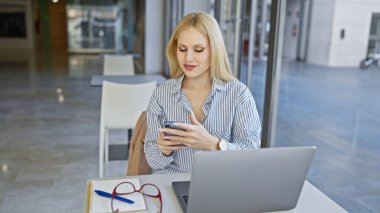 Doğal ışık ve şehir manzaralı modern bir kafede bilgisayarla çalışırken akıllı telefon kullanan sarışın bir kadın..